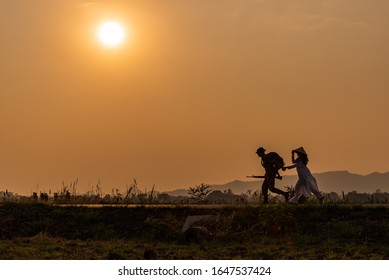 The Silhouette Of A Soldier And A Vietnamese Woman Running Away At Sunset,In The Field With Mountains Behind