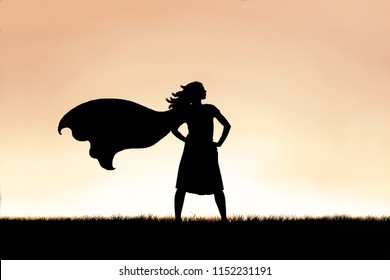 The silhouette of a strong, beautiful caped superhero woman stands isolated against a sunset in the sky background.