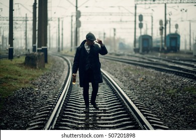 Silhouette Of Stylish Gangster Man With Bottle Whiskey Walking On Railway Road. England In 1920s Theme. Fashionable Brutal Confident Man. Atmospheric Moments