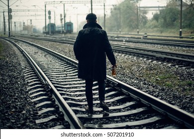 Silhouette Of Stylish Gangster Man With Bottle Whiskey Walking On Railway Road. England In 1920s Theme. Fashionable Brutal Confident Man. Atmospheric Moments