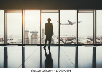 A silhouette of a successful woman entrepreneur standing indoors of a luxurious interior with a reflective marble floor, and a plane gaining altitude over an urban skyline outside a panoramic window