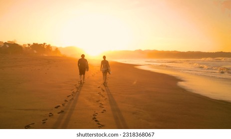 SILHOUETTE: Surf couple walking along gorgeous sandy beach towards golden sunset. Two friends on a holiday surf trip at Playa Venao. They are going surfing and riding waves in beautiful sunset light. - Powered by Shutterstock