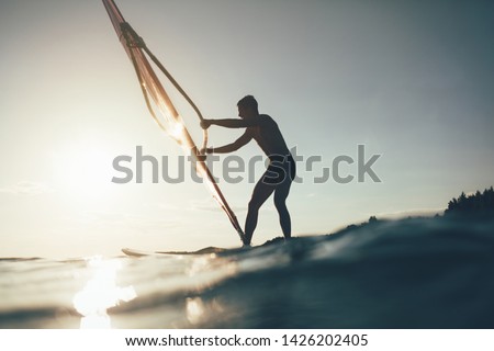 Silhouette of surfer balancing on windsurf board. Low angle splashing view of windsurfer. Windsurfing, sailing, water sports, extreme