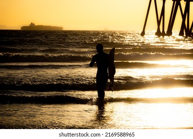 Silhouette Of A Surfer Walking On The Beach Carrying A Surfboard In Front Of The Pier At Huntington Beach, California At Sunset