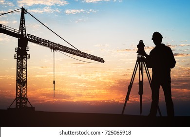 Silhouette Of A Surveyor Standing With Equipment Near Crane At Construction Site During Sunset