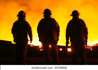 A Silhouette Of Three Fire Fighters Watching A Fire