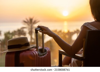 Silhouette of tourist woman with straw hat sitting on the armchair with suitcase on terrace and looking on sunset palm sea beach. Travel,vacation concept.Rear view - Powered by Shutterstock