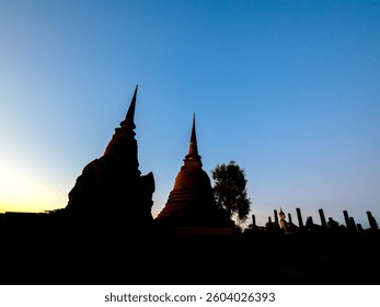 Silhouette of two ancient chedis (stupas) against a vibrant sunset sky at Sukhothai Historical Park, Thailand. 