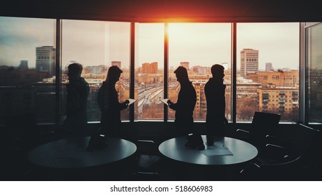 Silhouette Of Two Businessmen With Their Gadgets In Office Interior Of Skyscraper, Man On The Left With Digital Tablet And Man On The Right Talking On The Phone, Cityscape Outside, Strong Reflections