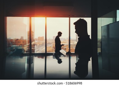Silhouette of two businessmen working with their gadgets in office interior of skyscraper, man in front with digital tablet and man behind with phone, cityscape outside, strong reflections - Powered by Shutterstock