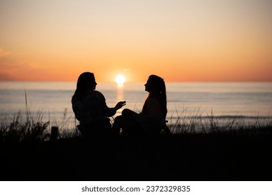 Silhouette of two female friends enjoying a conversation while sitting and watching the sunset on a sea beach. Back view
