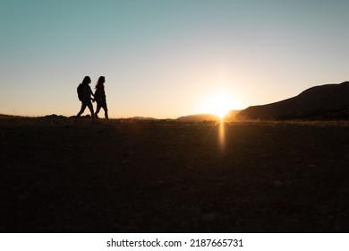 Silhouette Of Two Girls With Backpacks While Traveling In The Mountains At Sunset. Two People Walk Along The Mountain Range At Sunset. Travel With Freedom.