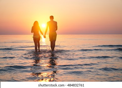 Silhouette Of Two Lovers Walking Hand To Hand On The Beach At Sunset Time - Young People Enjoying Vacation Holidays In Summer - Concept Of Love Relation And Romantic Moments - Original Sun Color Tones