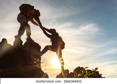 Silhouette Two Male Hikers Climbing Up Mountain Cliff And One Of Them Giving Helping Hand. People Helping And, Team Work Concept.