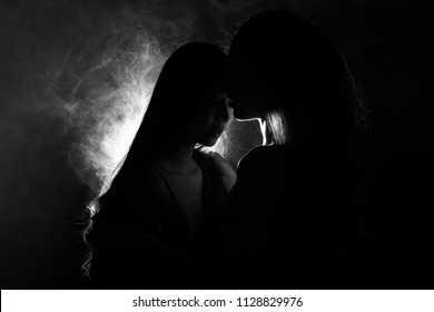 Silhouette of two sexy woman kissing holding in darkness through light and smoke, they have good feeling on first date flirt and want to love best friends forever, girls in long straight black hair