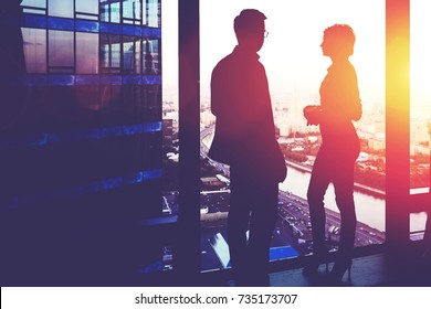 Silhouette Of A Two Successful Managing Directors Discuss Ideas After Meeting With Partners While Standing Near Big Office Window, Young Man And Woman Skilled Economists Relaxing After Conference