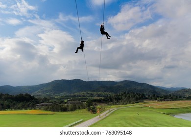 Silhouette Of Two Tourists Wearing Casual Clothing Are Playing Zip Line And Selfie With Big Mountain Background And Beautiful Cloud Blue Sky At Singha Adventure Park Chiang Rai, Thailand