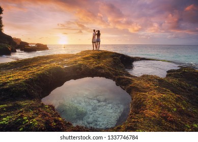 Silhouette of two young people looking like pair in love watching an amazing purple and orange coloured sunset over the sea with the salt water oceanic nature pool in the foreground