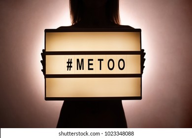 Silhouette Of Unknown Female In The Dark Covering Her Chest With #Metoo Hashtag Word On Glowing Lightbox. Me Too Light Box. Anti Sexism Protest Movement Against Inappropriate Behavior Towards Women.