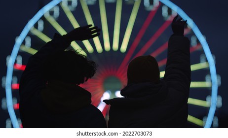 Silhouette Of Unknown Romantic Couple Jumping On Illuminated Ferris Wheel Background Outdoor. Unrecognizable Happy Man And Woman Waving Hands On Urban Street At Night. Urban Love Concept.