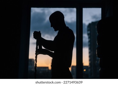 Silhouette of unrecognizable boxer male wrapping defense boxing tape around wrist standing on background of window on evening during sunset. Strong fighter preparing to wear gloves in fitness center. - Powered by Shutterstock