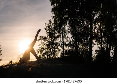 Silhouette Of Unrecognizable Woman Practicing Three-Legged Downward-Facing Dog Yoga Asana Pose In Evening At Sunset. Girl Performing Eka Pada Adho Mukha Svanasana Posture Outside. 