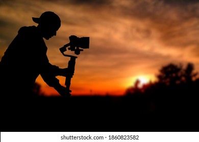 Silhouette of videographer is filming with cinema gimbal video dslr at sunset , professional video, videographer in events. Cinema lens on gimbal. Medium shot from right side. Film or cameraman school - Shutterstock ID 1860823582