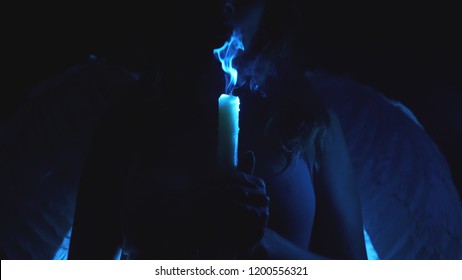 silhouette of a woman with angel wings, holding a candle in her hands. blow off the flame of the candle, the smoke from the wick of the candle in the dark. against the background of angelic wings.