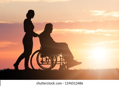 Silhouette Of Woman Assisting Her Disabled Father On Wheelchair In Park At Sunset