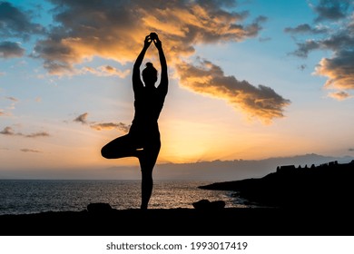 The silhouette of a woman doing yoga stretches on the beach during sunset, holding an apple above her head