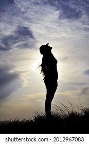 Silhouette of a woman in a full-length cap against a blue sky on top of a hill among tall grass, looking up and making a wish, copy space.