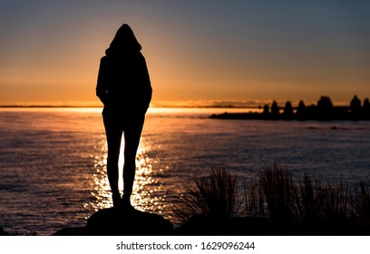 Silhouette of woman in hoodie standing on lake shore in front of sunrise. - Powered by Shutterstock