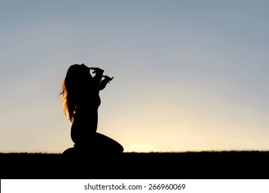 A silhouette of a woman kneeling down with her hands in the air, praying, thanking, and surrendering to God.