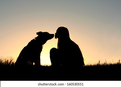 a silhouette of a woman with long blonde hair is sitting outside in the grass, kissing her large German Shepherd Mix dog at sunset - Powered by Shutterstock