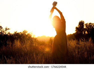 Silhouette of a woman with long natural hair in a long mustard-colored cotton dress picks wild flowers against the background of the sunset. Natural beauty, privacy in nature, women's day. - Powered by Shutterstock