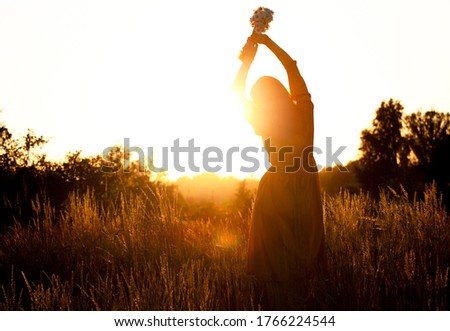 Silhouette of a woman with long natural hair in a long mustard-colored cotton dress picks wild flowers against the background of the sunset. Natural beauty, privacy in nature, women's day.