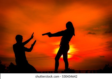 Silhouette Woman Pointing A Gun. Mafia Woman Pointing At The Man On The Sunset Background.Silhouette Concept