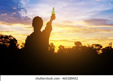 Silhouette Woman Raised Hands Holding A Green Beer Bottle On The Sunset Sky,