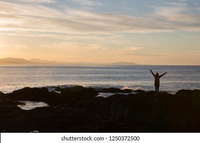 Silhouette Of Woman Raising Hands In Praise At A Rocky Beach During Sunset With Waves Breaking