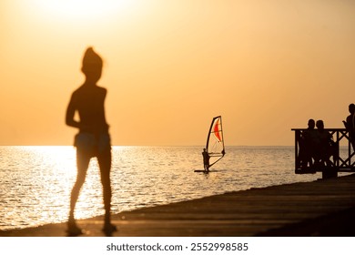 Silhouette of a woman standing on a pier at sunset, watching a windsurfer gliding across the sea - Powered by Shutterstock