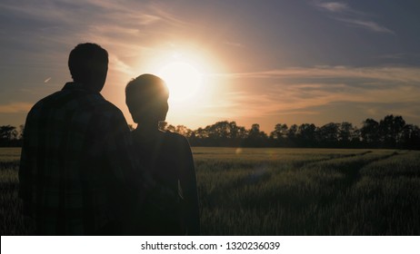 Silhouette woman standing at the sunset rear back view The man approaches and hugs the waist Couple resting enjoy beautiful landscape at sundown. Field with young plant and trees. Exiting paysage at