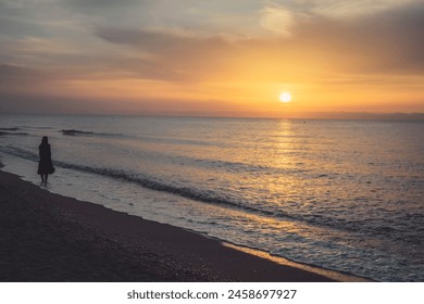 The silhouette of a woman walking along the beach at dawn, the sky painted in orange tones and the calm water. - Powered by Shutterstock