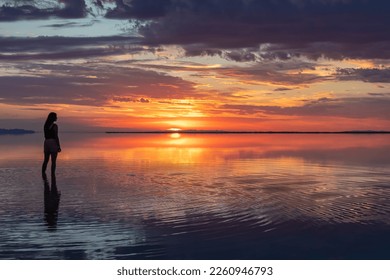 Silhouette of woman walking into the sunrise of lake Bonneville Salt Flats, Wendover, Western Utah, USA, America. Dreamy red colored clouds mirroring on the water surface creating romantic atmosphere - Powered by Shutterstock