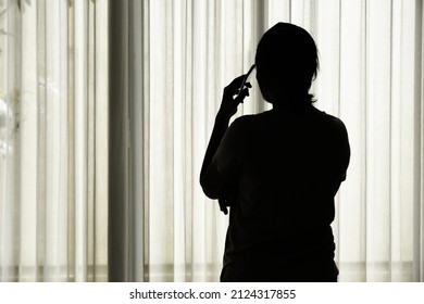 Silhouette Of A Woman Wearing Mask In Her Quarantine During Corona Pandemic, Making A Phone Call From Behind The Window Curtain