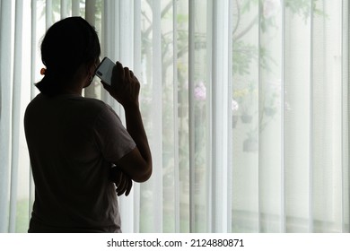 Silhouette of a woman who is quarantined at home due to covid-19. She is standing by the window making a phone call.