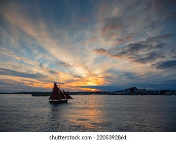 Silhouette of wooden sail boat going into harbor. Dark and dramatic sunset sky. Galway hooker wooden boat type. Galway city, Ireland. Water sport and hobby.