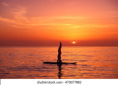 Silhouette of a yoga trainer balancing on the water on the paddle board over beautiful orange sunset background, standing on the head, doing Sirshasana on the beach