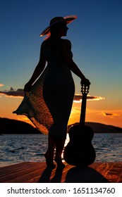 Silhouette young adult female standing pier near to the sea in long dress   hat looking the right  holding guitar orange sunset