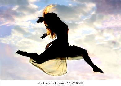 A silhouette of a young, attractive, dancing woman jumps through the air, with a beautiful cloudy sunset in the background outside.