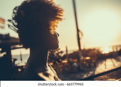 Silhouette of young Brazilian girl with curly afro hair illuminated by orange light looking on coastline while standing on embankment on windy summer evening, with beach cafe in defocused background - Powered by Shutterstock